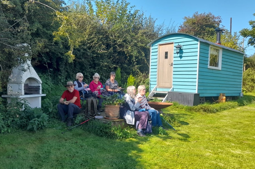 group members resting in the shade near a wooden caravan and wood burner